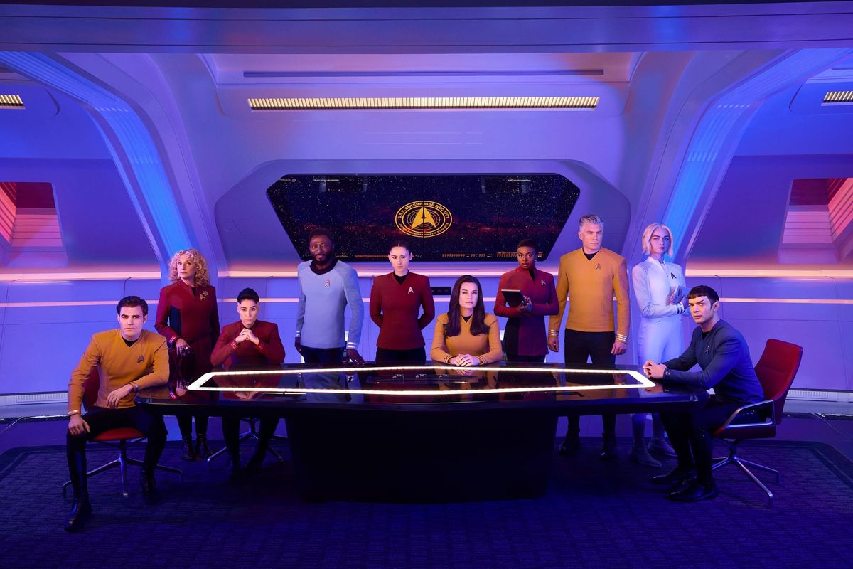 The cast of Star Trek: Strange New Worlds sits around a glowing table in a starship setting, wearing their Starfleet uniforms.