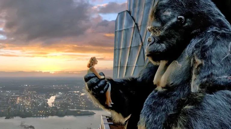 An enormous gorilla holds a small, white blonde woman in his paw. They sit high above the city, at the top of a skyscraper.