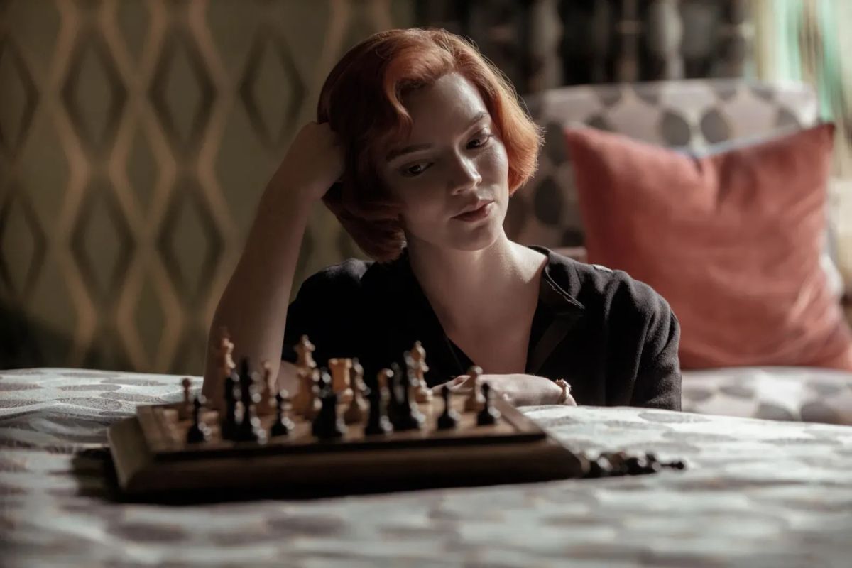 A woman with a redheaded bob regards a chessboard that has been set on a bed. She is in '60s fashion.