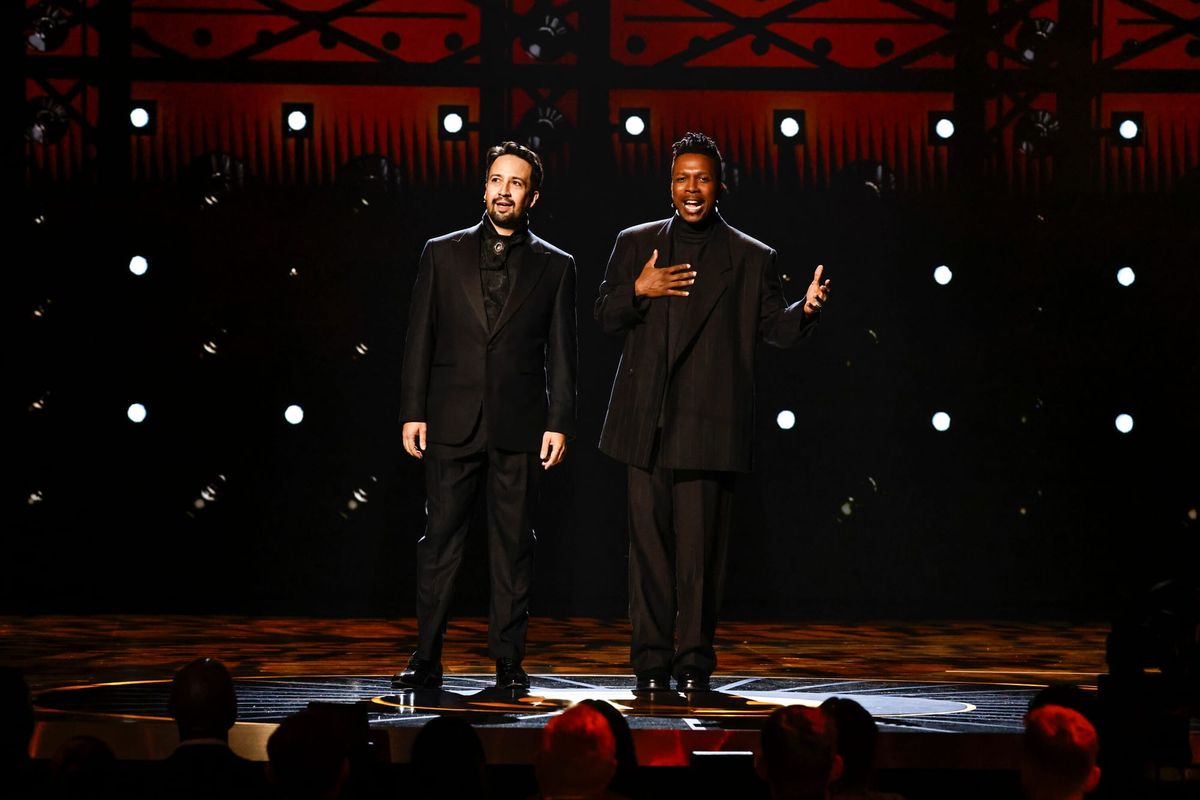 Lin-Manuel Miranda and Leslie Odom, Jr., both in all black suits, perform on a large stage.