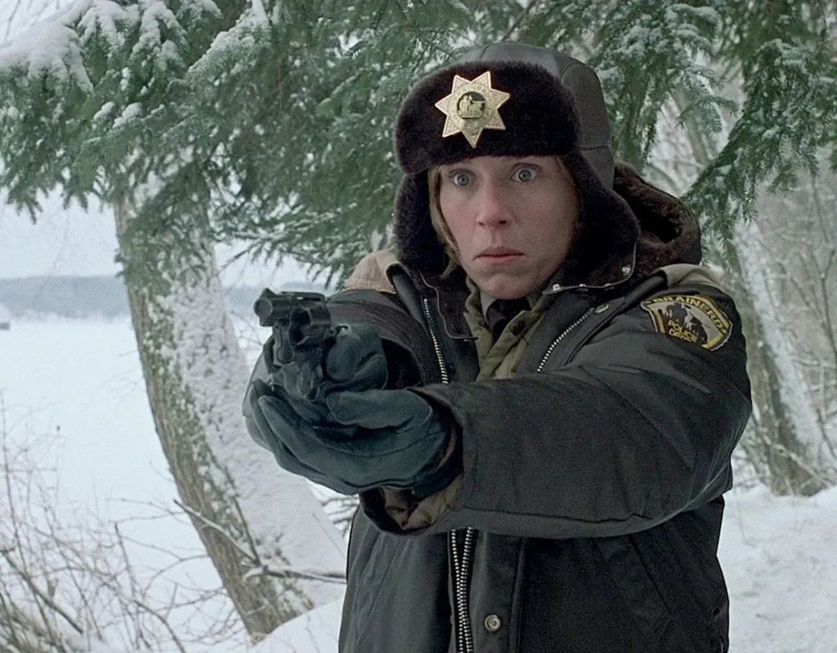 A white woman in a police uniform pulls a gun. Everything is snowy, and she is dressed very warmly.