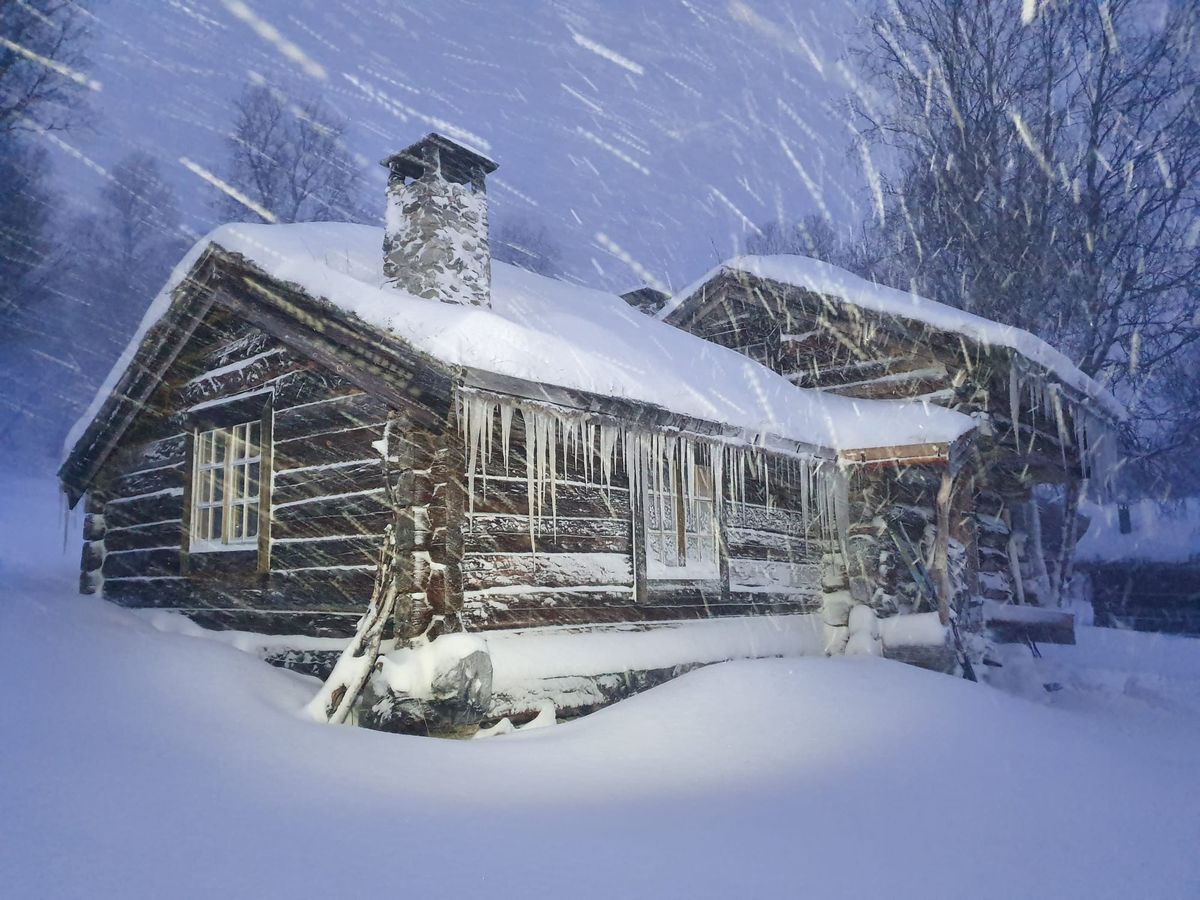 A cabin in the midst of a terrible snowstorm. It is covered in snow, and icicles hang all over from it.