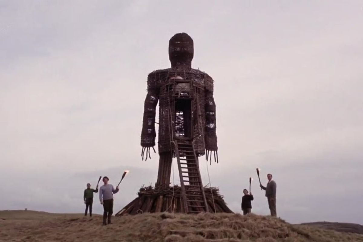 An enormous wicker man stands on a hilltop, surrounded by four smaller figures, all real humans bearing torches.