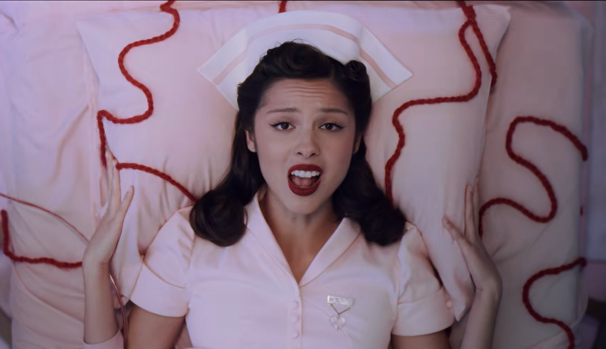 Olivia Rodrigo, wearing a nurse's outfit, lays on a hospital bed, long trails of red yarn running from her body like blood.