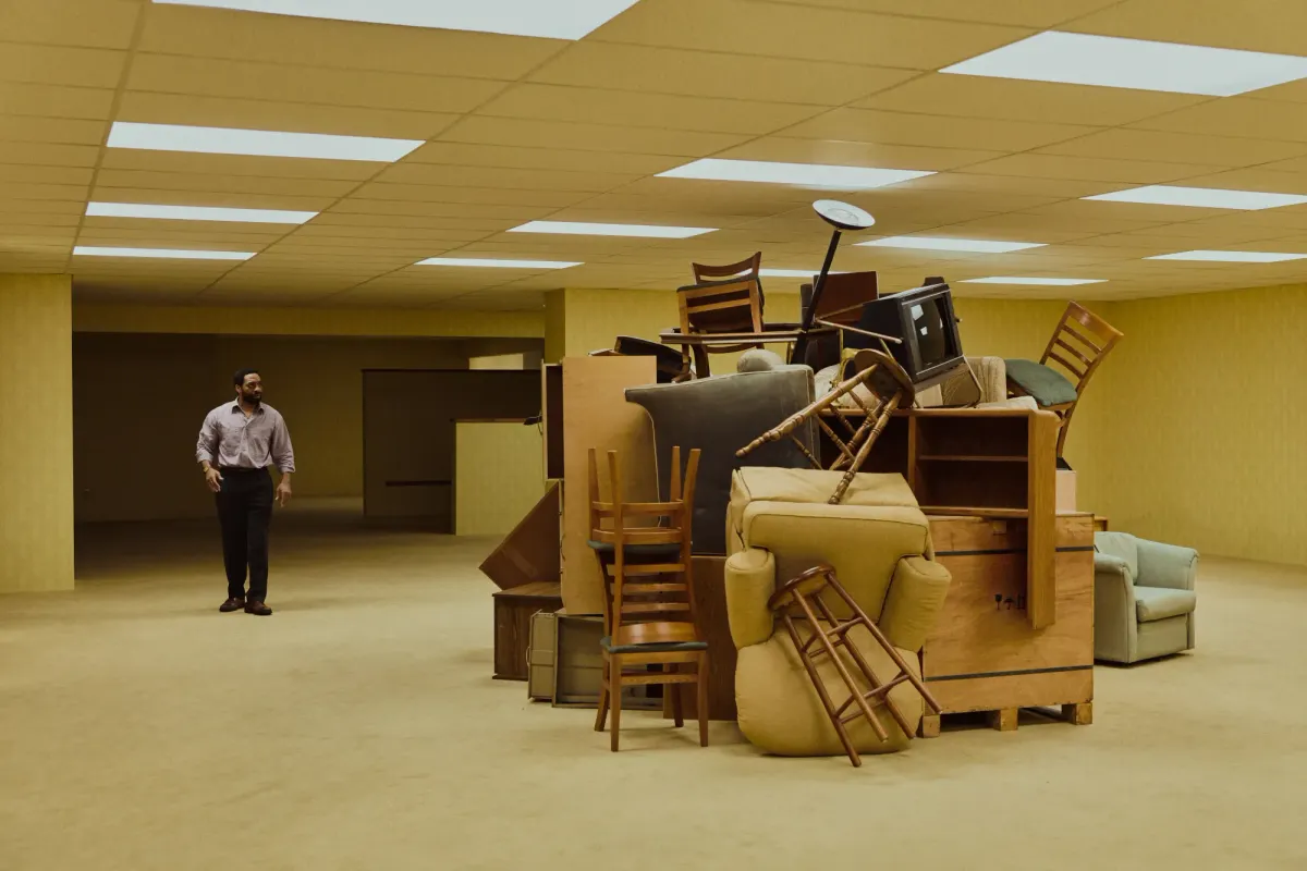 A man walk through a non-descript, yellow room with an enormous pile of furniture at its cent