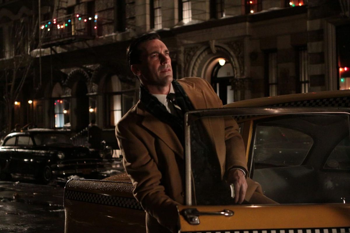 Don Draper gets out of a taxi, looking up at an apartment building. Christmas lights abound, and he wears a brown coat.