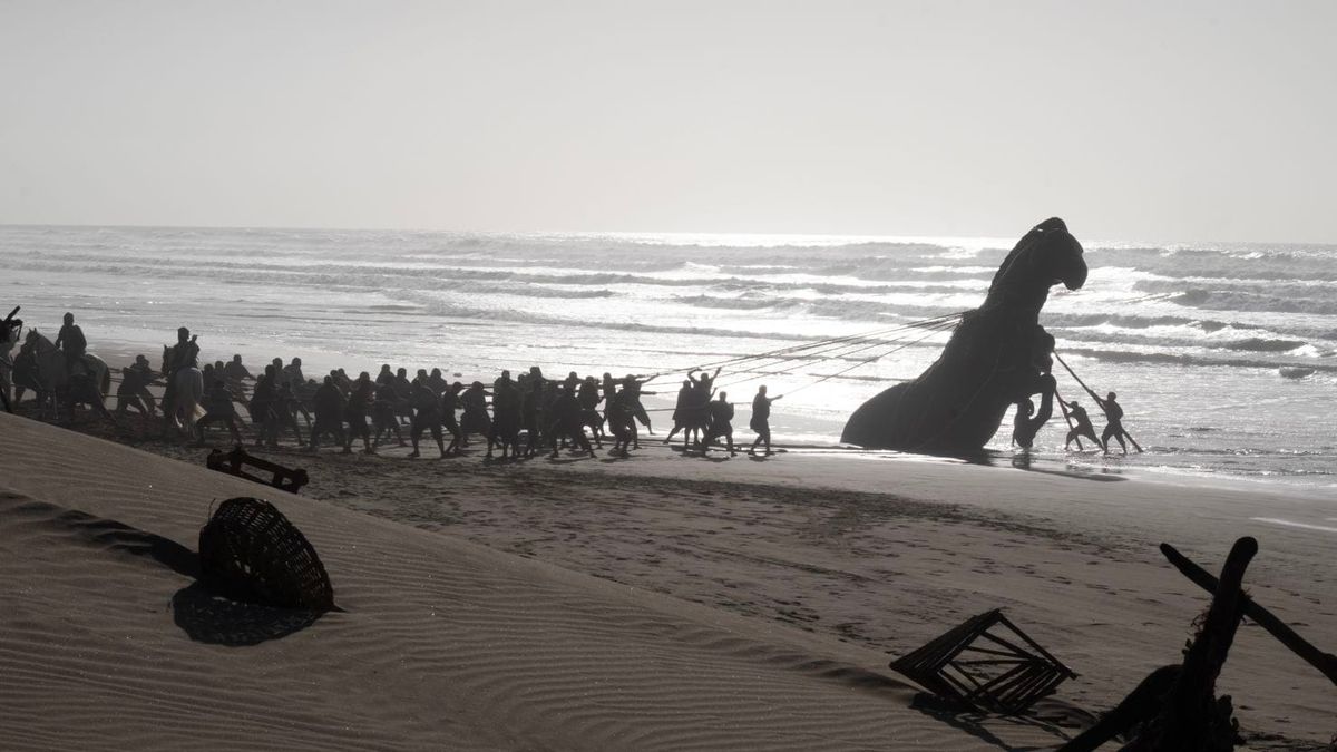 An army of men tug an enormous wooden horse out of the sand, waves crashing in the background.