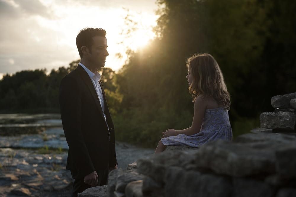 Kevin (Justin Theroux, in a lovely suit) looks at a young girl, who sits at the edge of a well. The risisun pokes through the trees behind them.