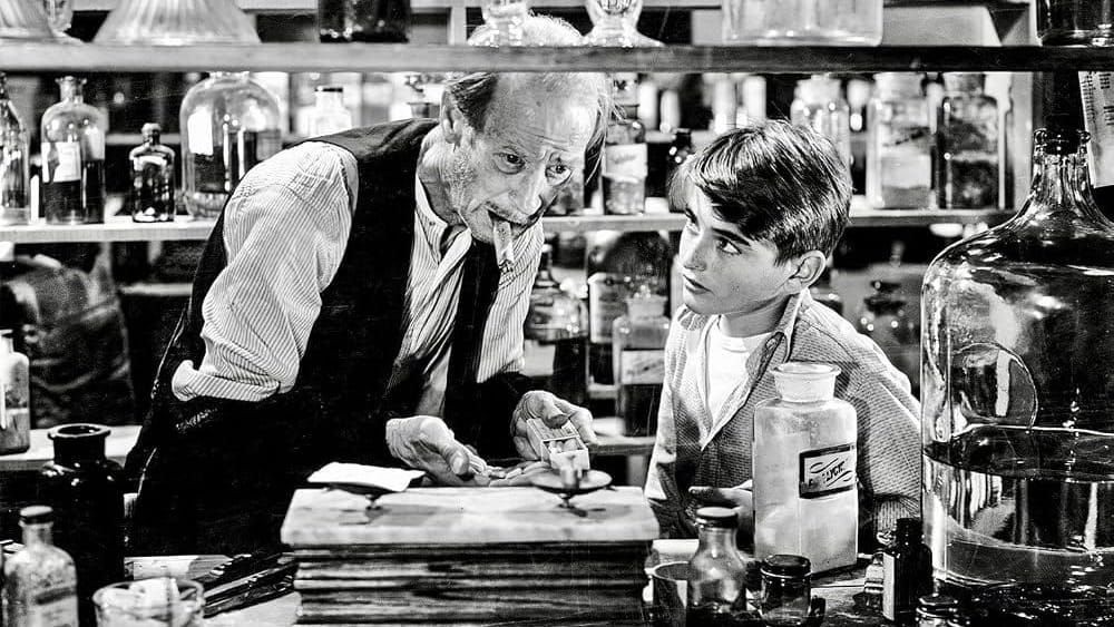 Mr. Gower, an old man, takes in what the young George Bailey tells him. They are surrounded by drug and pill bottles. The scene is in black and white.