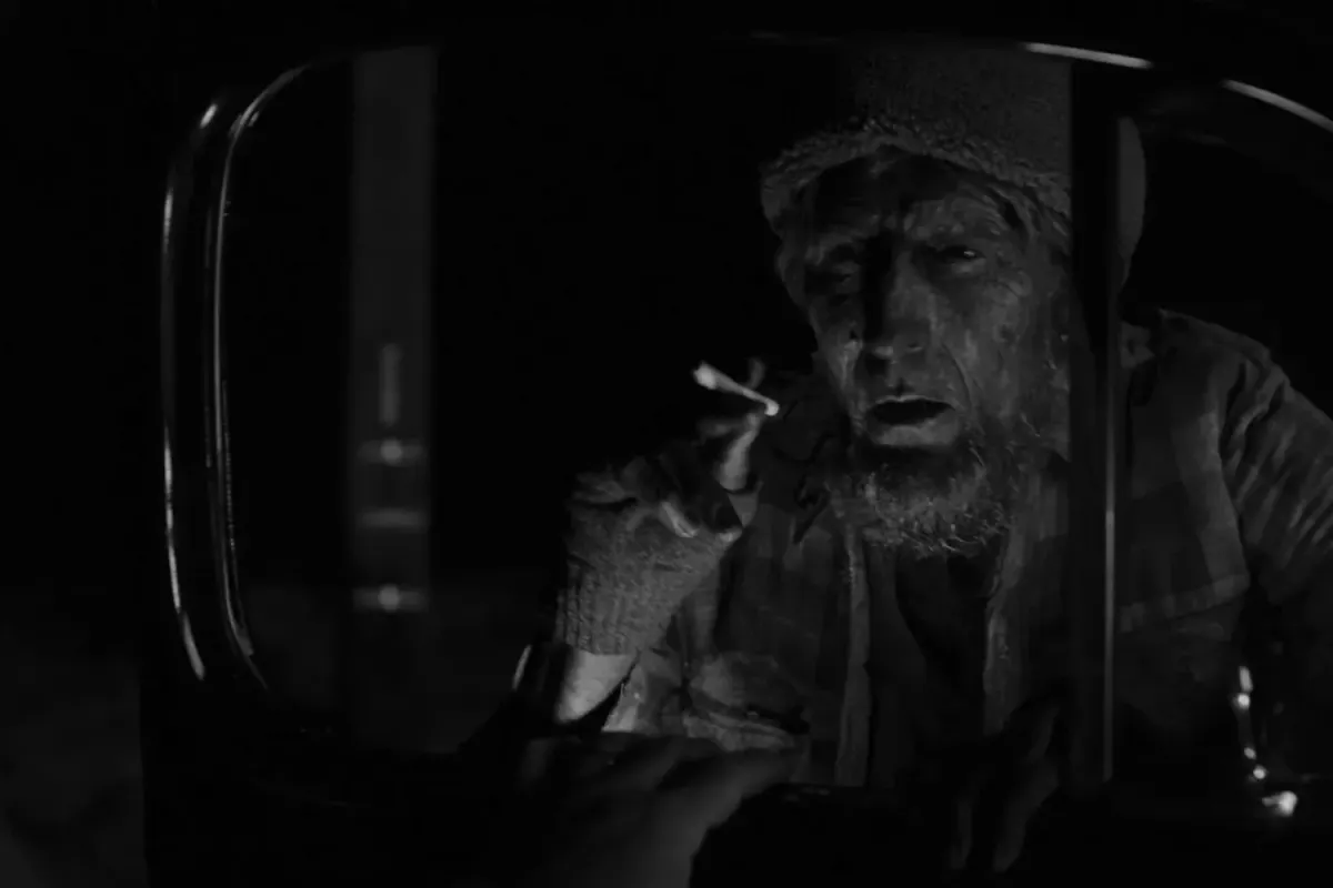 A ghostly man with a beard, dressed for the outdoors, leans into a car window, cigarette held out for a light. The image is in black and white
