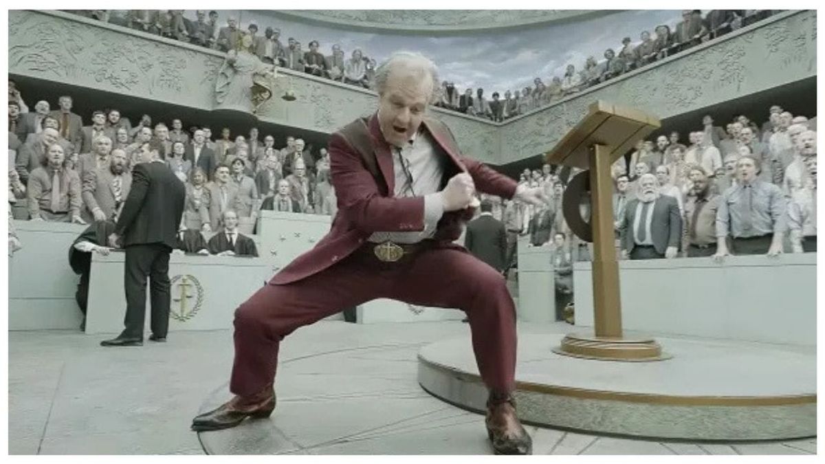 Digger Rockwell, in a burgundy suit, gets real excited as he addresses a large room full of people clothed in various shades of beige.