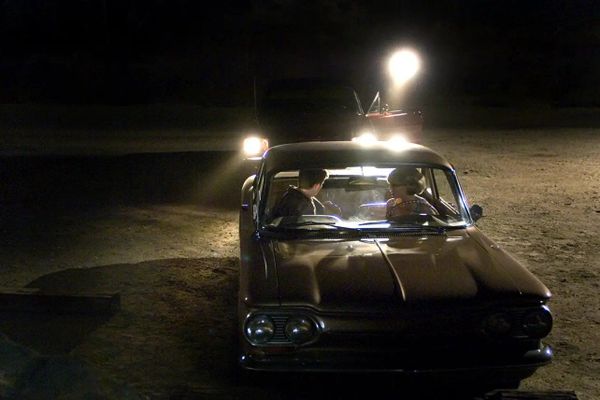 Two people in a car look back at a car parked behind them. That car's driver shines a flashlight at them.