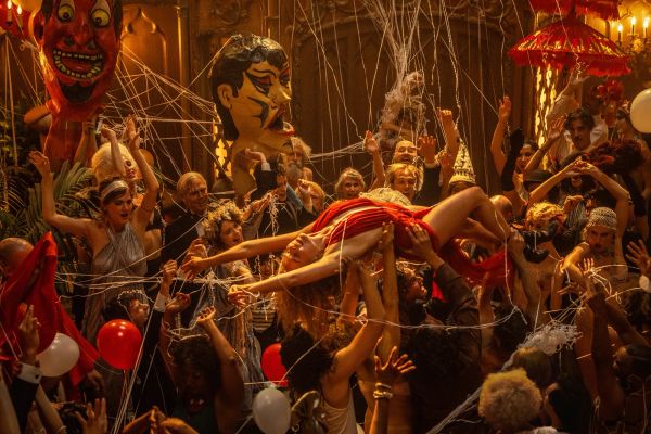 Margot Robbie, in a red dress, rides atop a throng of people, borne up by their hands, at a massive party.