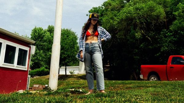 Katie Crutchfield, aka Waxahatchee, stands in the middle of greenery in a shirt over a red bra. She wears a ball cap and 
