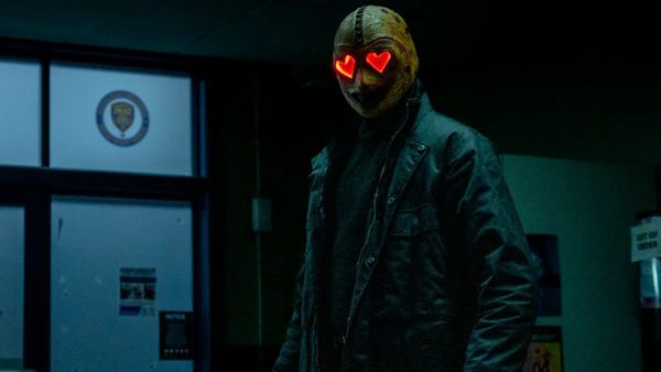 The Heart Eyes Killer stands in a darkened room. Their red heart eyes glow in their otherwise yellow mask.