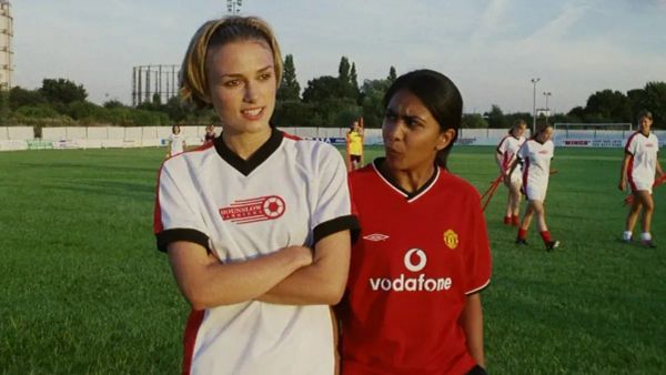 Two young women walk across a soccer pitch in their jerseys. One is tall and white, the other shorter and of Indian descent.