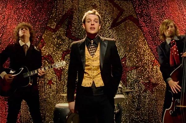 The Killers perform Mr. Brightside in the music video. They are wearing dapper suits. Behind them is a sparkly backdrop.
