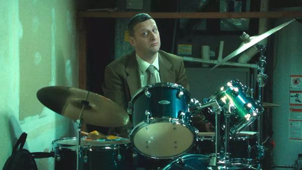 Tim Robinson, clad in a deeply tan suit, sits behind a sweet drum set.