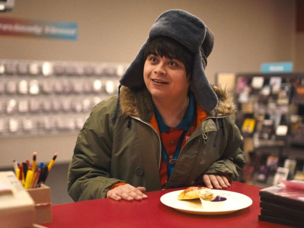 A white teenage boy wearing a ridiculous hat with earflaps is in a video store. A piece of pizza sits before him.