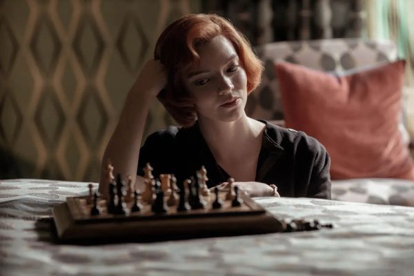 A woman with a redheaded bob regards a chessboard that has been set on a bed. She is in '60s fashion.