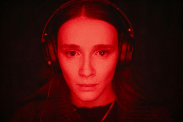A young with woman with brown eyes, wearing headphones, stares straight at something on a screen. She is bathed in red light.