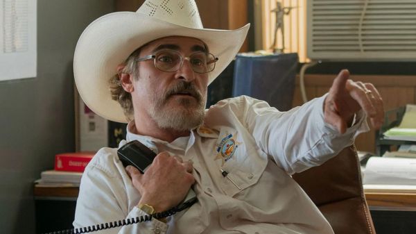 Joaquin Phoenix, in a white cowboy hat and white sheriff's shirt, holds a radio and points at someone off-camera.