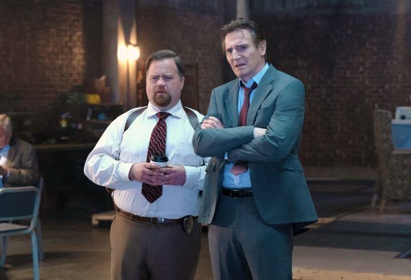 Paul Walter Hauser and Liam Neeson star in The Naked Gun. They're in a non-descript room of brick. Hauser clutches coffee.