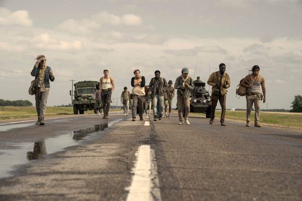 A group of young men walk down a long stretch of highway. They look absolutely exhausted. Men in military vehicles follow.