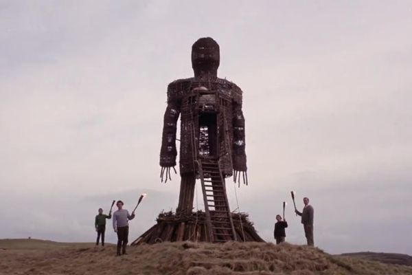 An enormous wicker man stands on a hilltop, surrounded by four smaller figures, all real humans bearing torches.