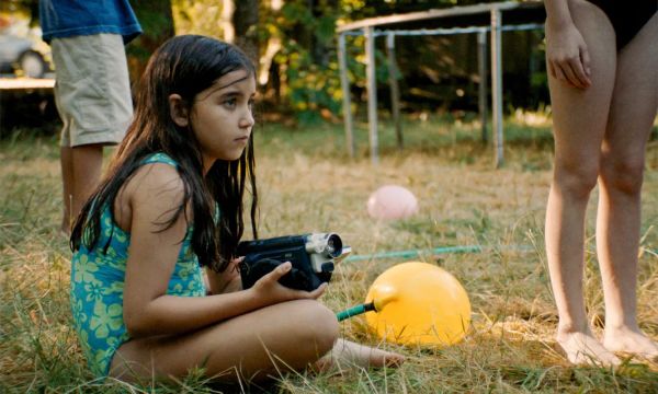 A young girl with long dark hair in a blue and green swimsuit films something on a handheld camcorder.