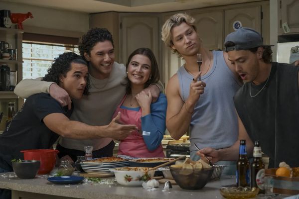 A group of young men gather around a counter where they are preparing Thanksgiving food. One of them has his arm around a young woman.