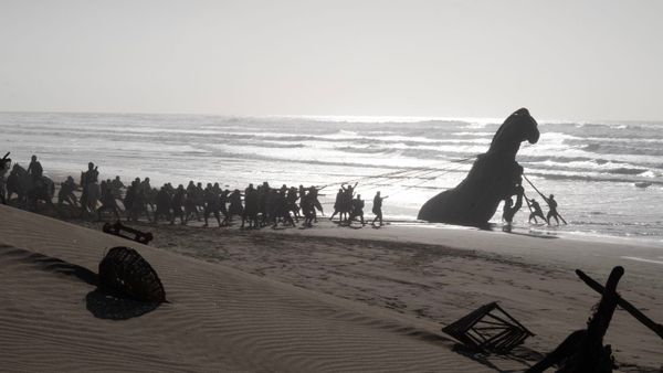An army of men tug an enormous wooden horse out of the sand, waves crashing in the background.