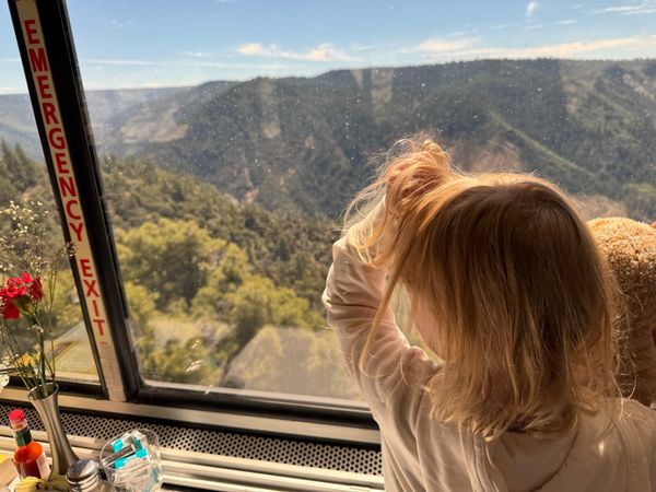 A tiny child looks out the window of a train as it rolls along a high mountain ridge.