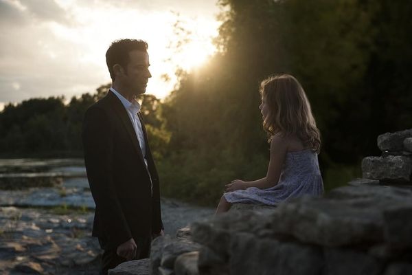 Kevin (Justin Theroux, in a lovely suit) looks at a young girl, who sits at the edge of a well. The risisun pokes through the trees behind them.