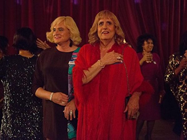 Marcie and Maura, dressed in vintage '90s fashion, enter a cross-dressing event with great joy.