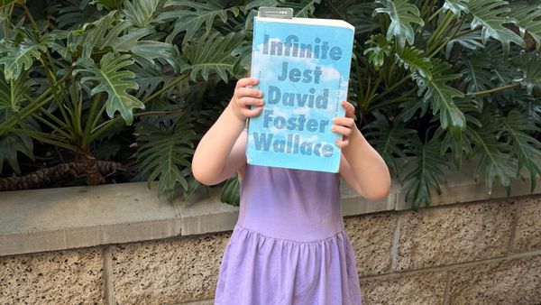 A small child in a purple dress holds a copy of Infinite Jest in front of their face. They stand in front of a bunch of dark green plants and a brick wall.