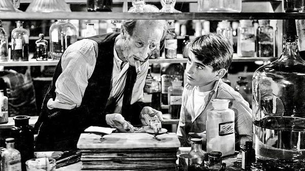 Mr. Gower, an old man, takes in what the young George Bailey tells him. They are surrounded by drug and pill bottles. The scene is in black and white.