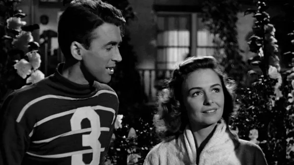 George Bailey (Jimmy Stewart), clad in a vintage football jersey, gazes longingly upon Mary Hatch (Donna Reed), wearing a bathrobe. She gazes out of the frame. They're surrounded by flowers.