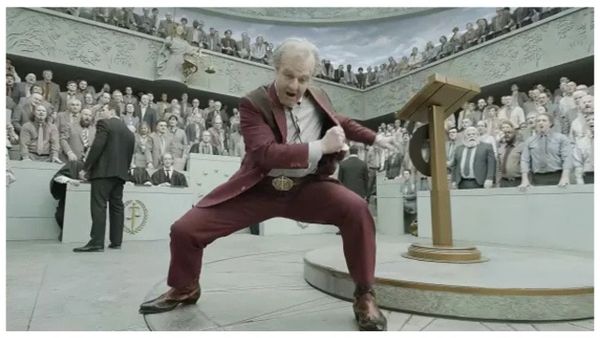 Digger Rockwell, in a burgundy suit, gets real excited as he addresses a large room full of people clothed in various shades of beige.