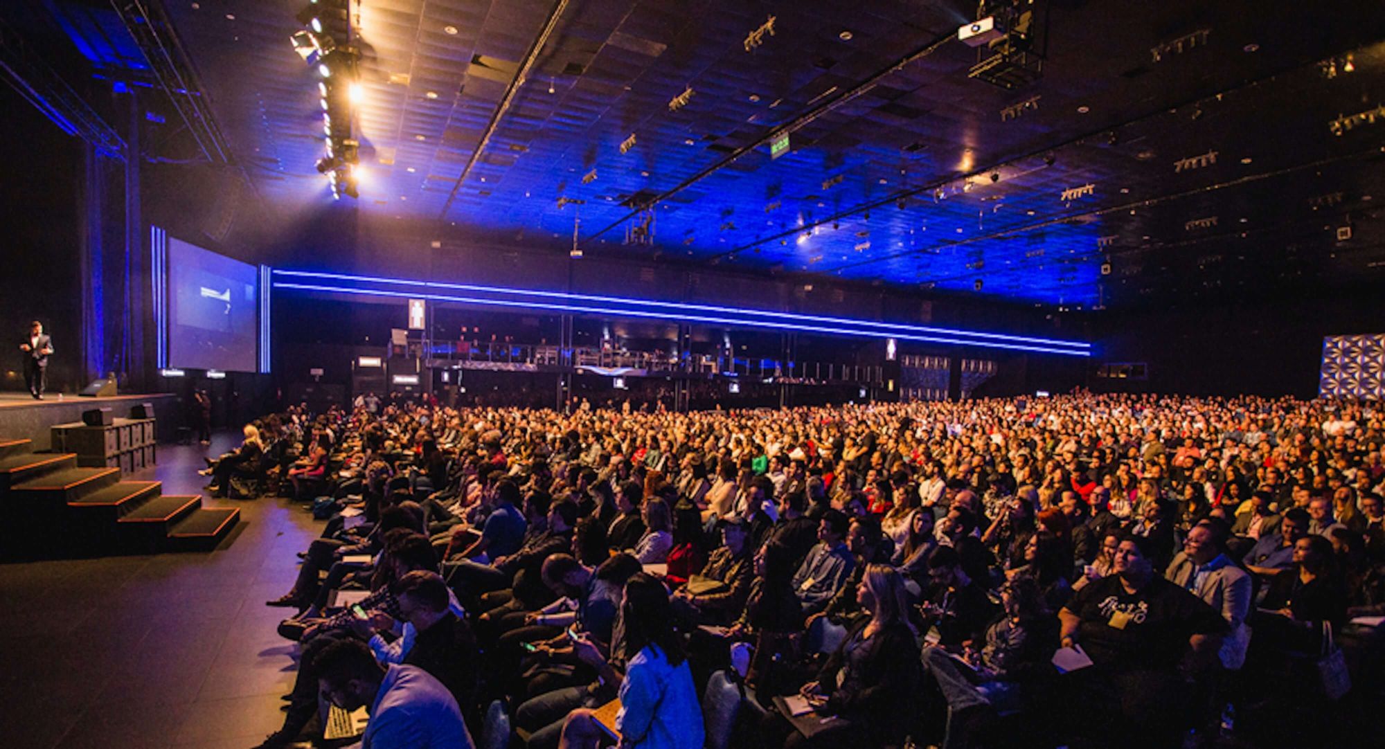 Seated audience (100+ people) with man stood on stage presenting.