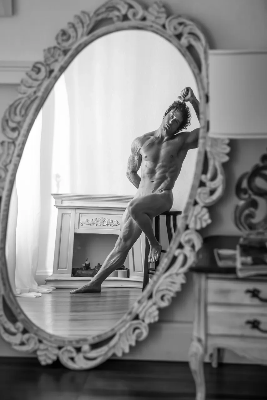 Artistic nude male figure reflected in an ornate mirror, sitting cross-legged with one arm behind and the other in hair, inspired by classical Greek art.