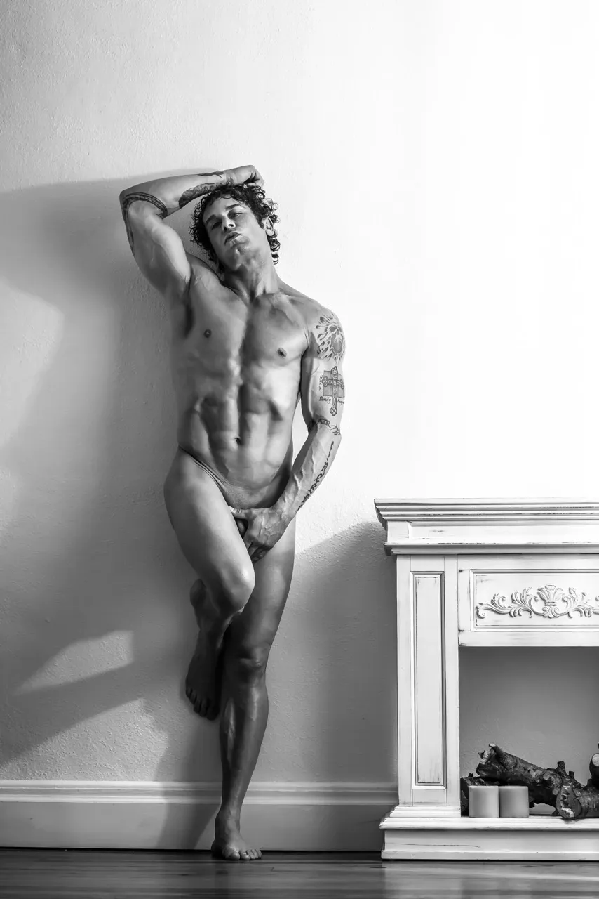 Nude man standing against a wall with legs crossed and hand covering genitals, echoing a modest classical statue pose with a modern, powerful twist.