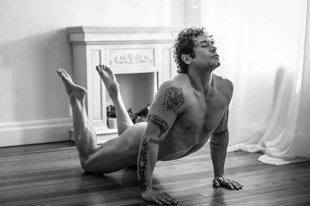Athletic nude man in an upward arched yoga pose on the floor, echoing Greek statue form with legs lifted and chest extended, lit by soft natural light.