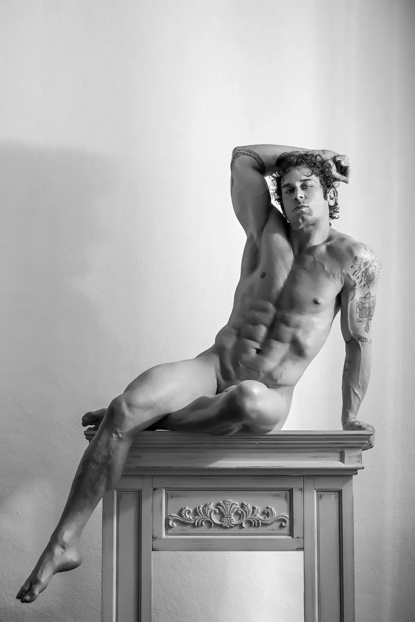 Masculine nude model posing atop a vintage mantel, legs crossed elegantly with one arm overhead, styled like a reclining ancient sculpture.