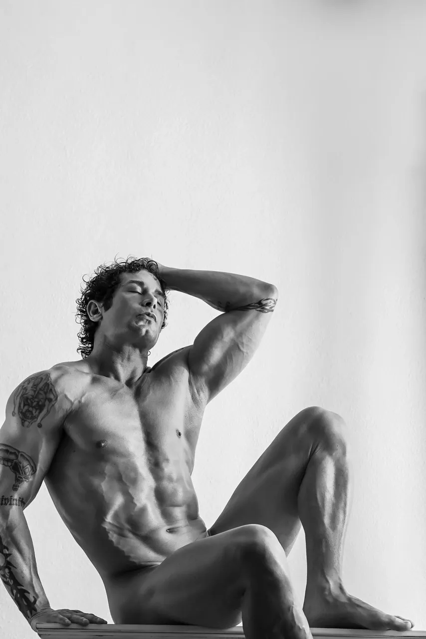 Close-up of a male nude model in a sensual sitting pose, abs and torso highlighted by directional light, evoking ecstasy and classical emotion.