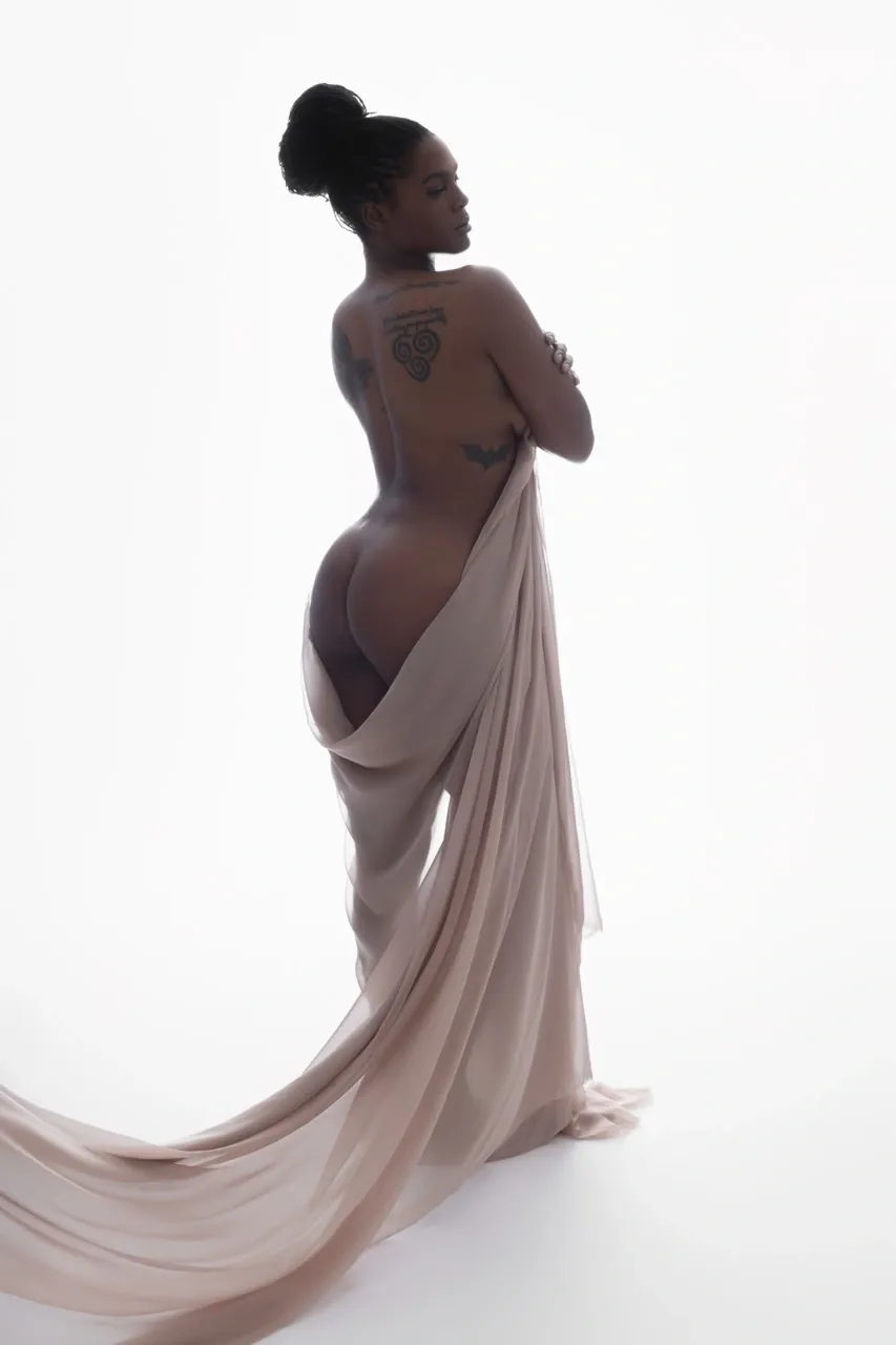 Nude fine-art portrait of woman draped in sheer fabric, back turned to camera with tattoos visible and high-key lighting.