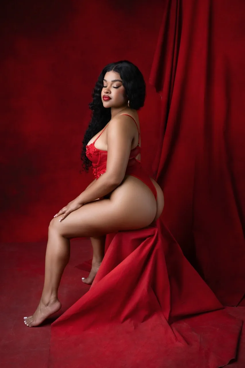 Curvy Black woman in red lingerie posing confidently on draped red fabric in a bold monochromatic boudoir studio with rich red backdrop.