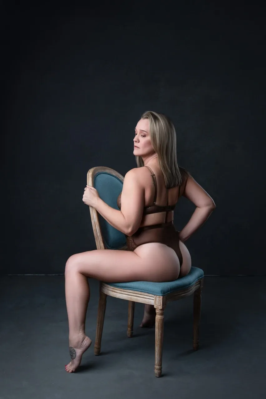 Boudoir portrait of a woman sitting backwards on a chair in sheer lingerie, highlighting her curves and booty with soft studio lighting against a dark blue background.
