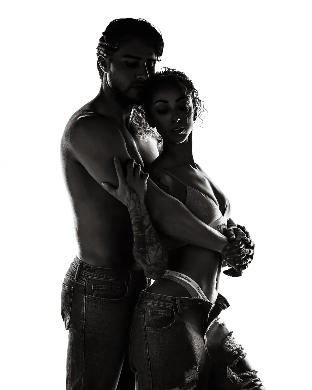 Black and white couples boudoir portrait featuring a shirtless man embracing his partner from behind as she rests against his chest in lingerie and jeans. Their intertwined hands, closed eyes, and relaxed body positioning emphasize romantic tension, trust, and sensual connection.