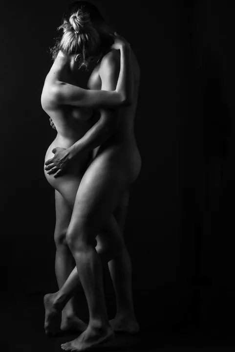 Black and white artistic nude couples boudoir photography capturing two partners standing in a full-body embrace, with one woman's leg crossed in front of her partner's body. Their intertwined arms, gentle touch, and natural body curves create a sensual composition focused on closeness and intimacy.